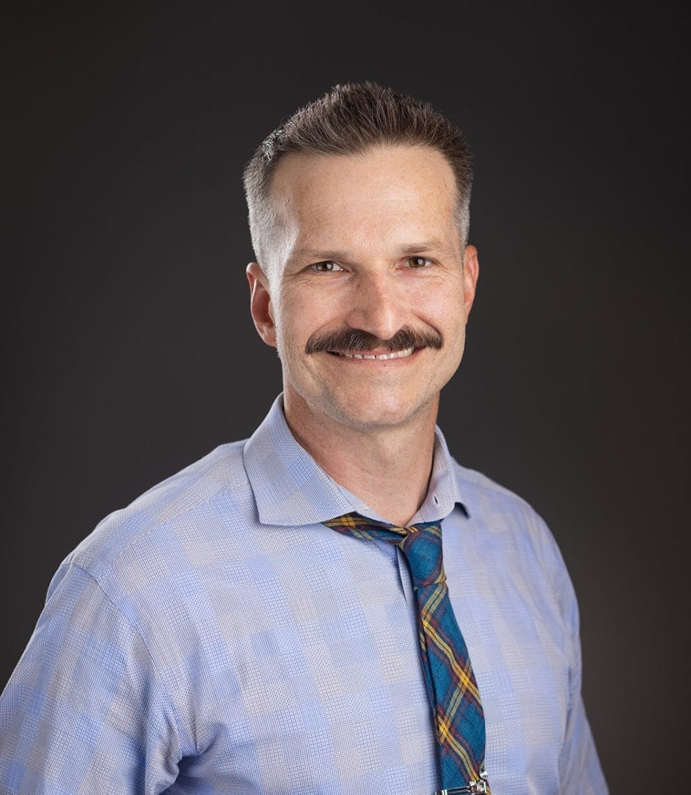 A man with short brown hair and a mustache, wearing a light blue checkered shirt and a plaid tie, smiles at the camera against a dark background.