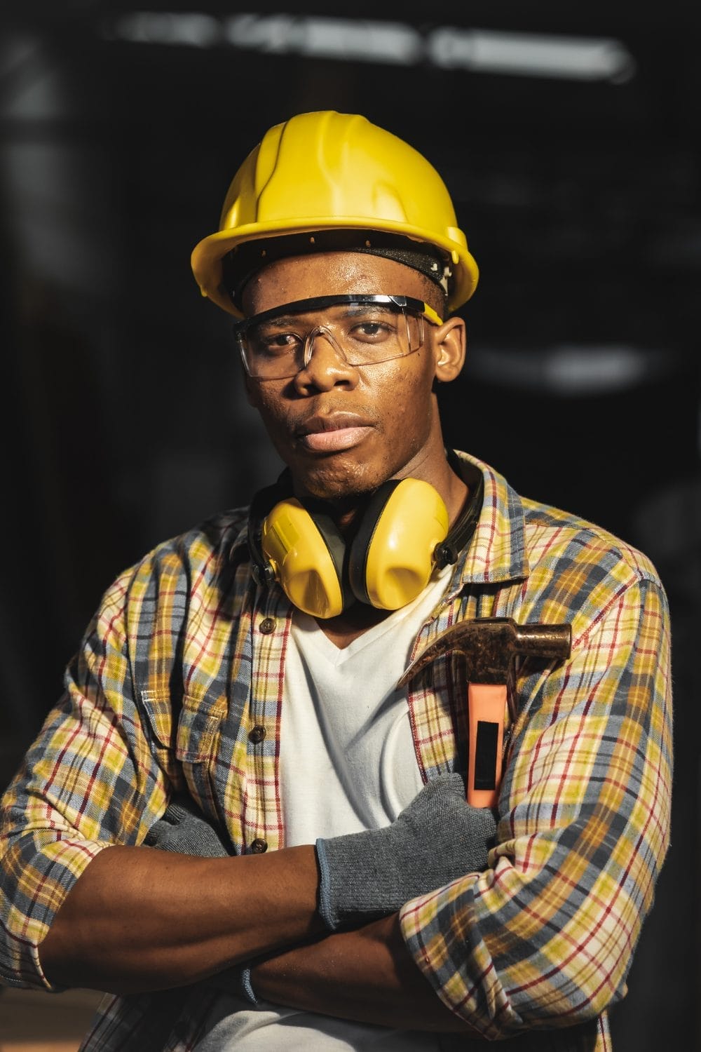 A construction worker in a yellow hard hat, safety goggles, and gloves stands with arms crossed, holding a hammer. This YouthBuild Salesforce NPSP implementation case study captures confidence and readiness on the job site.