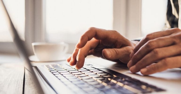 Hands typing on a laptop keyboard, with a cup of coffee in the background. The scene is lit by natural light coming from a nearby window.