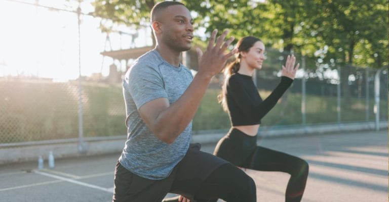 Two people are exercising outdoors on a sunny day. They are performing lunges on a paved surface next to a chain-link fence, with trees in the background. The man wears a gray shirt, and the woman wears a black outfit.