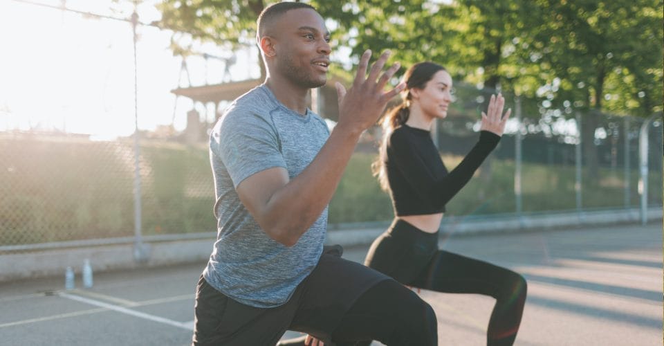 Two people are exercising outdoors on a sunny day. They are performing lunges on a paved surface next to a chain-link fence, with trees in the background. The man wears a gray shirt, and the woman wears a black outfit.
