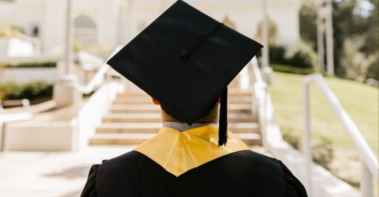 A graduate in a black cap and gown with a yellow stole stands facing a set of stairs outdoors. The background is blurred, showing greenery and part of a building. The focus is on the back of the graduate.