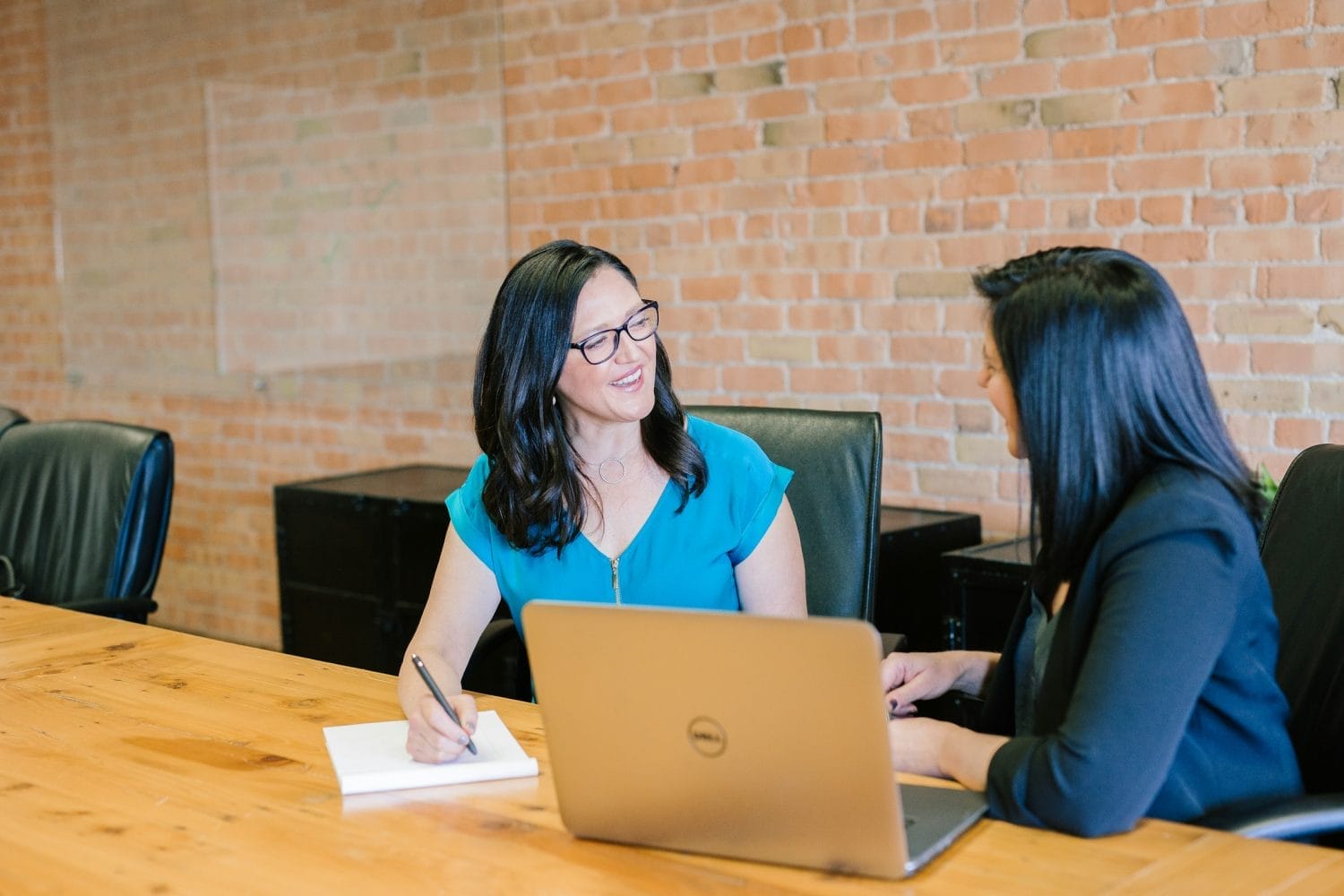 Two women sit at a conference table in an office. One is writing in a notebook while the other works on a laptop. Both are smiling and appear to be having a friendly conversation.