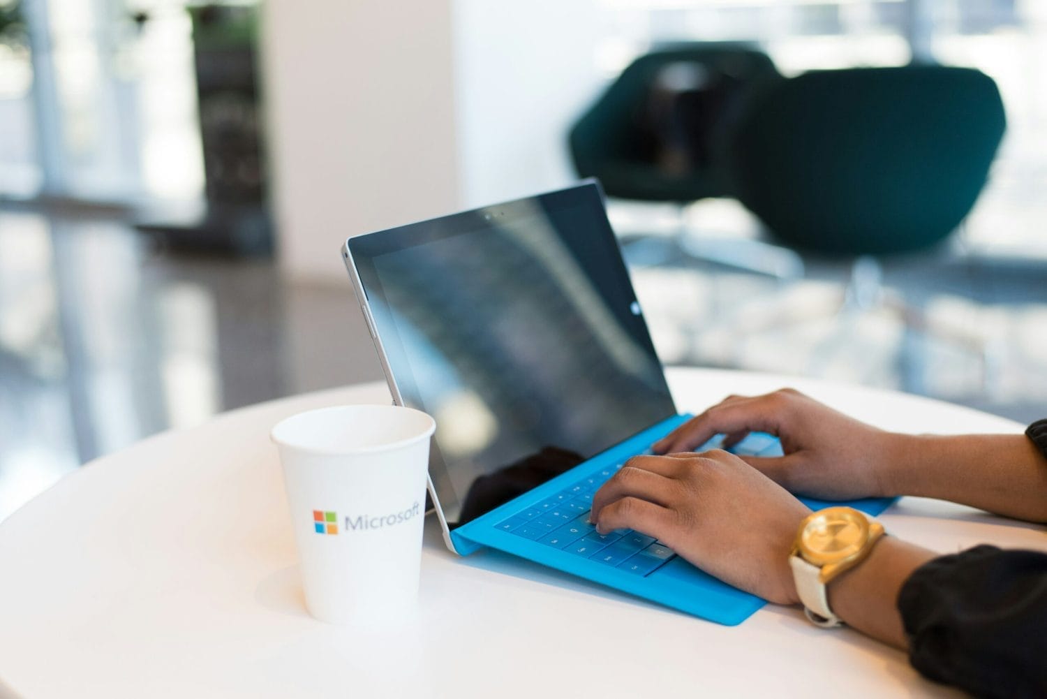 A person types on a Microsoft Surface tablet with a blue keyboard at a white table. A white cup with the Microsoft logo is next to the device, and the background is a modern, blurred office space.