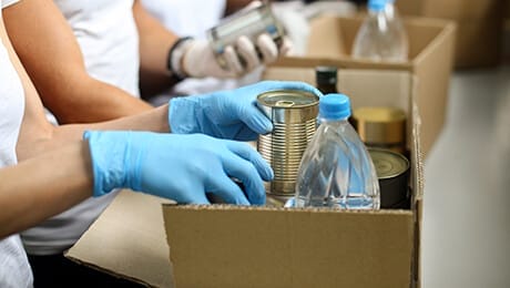 People wearing gloves pack canned goods and bottled water into a cardboard box, suggesting a food donation or relief effort.