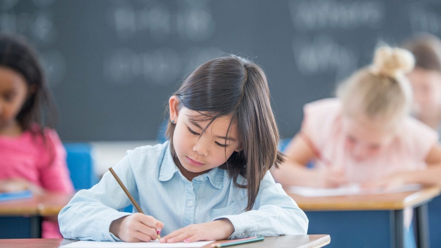 A young girl with straight dark hair sits at a desk in a classroom, focused on writing with a pencil. Other children are sitting and writing at desks nearby, with a chalkboard visible in the background.