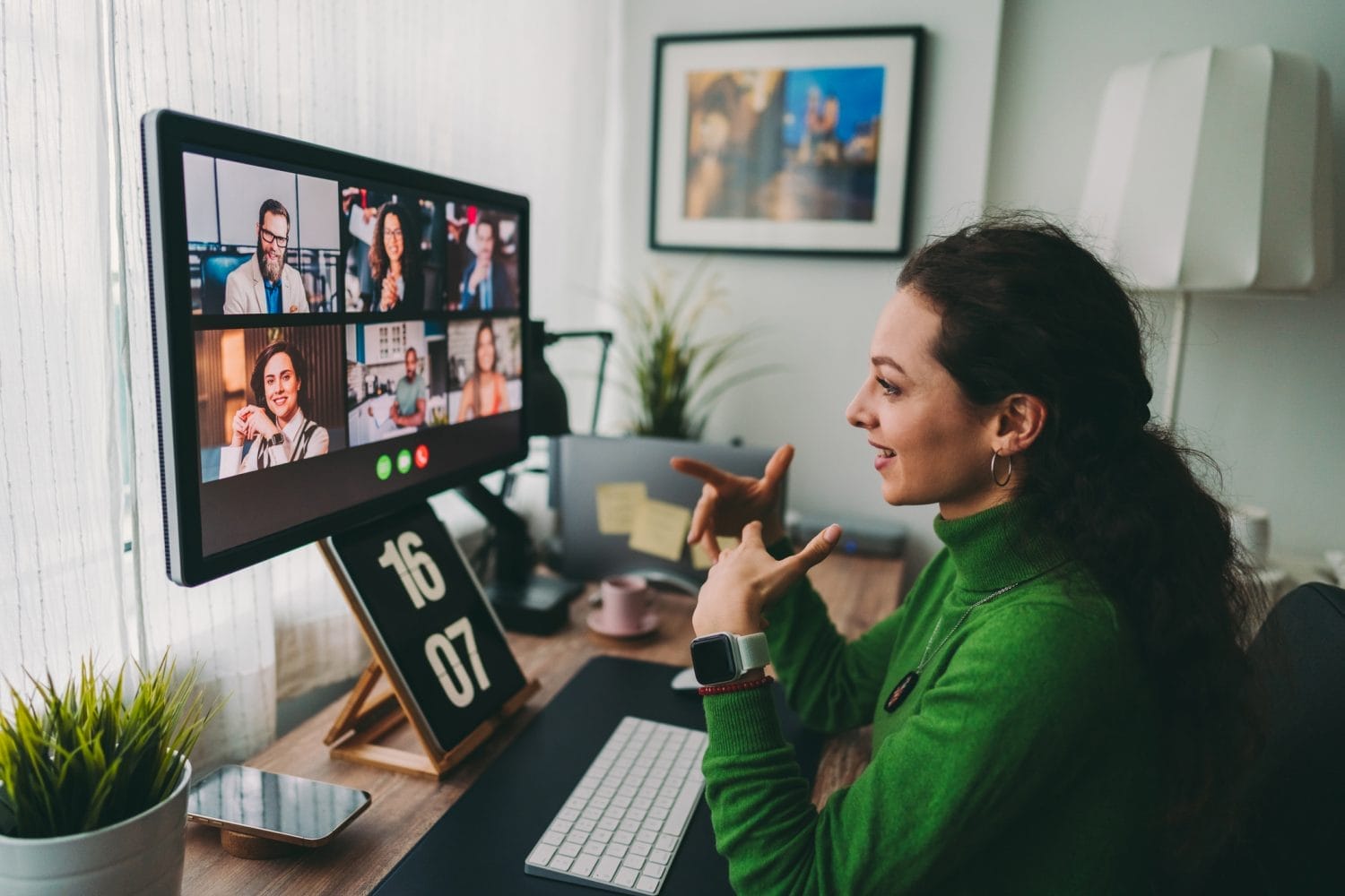 A woman in a green sweater sits at a desk, participating in a video conference with multiple people displayed on her computer screen. She gestures with her hands while speaking.
