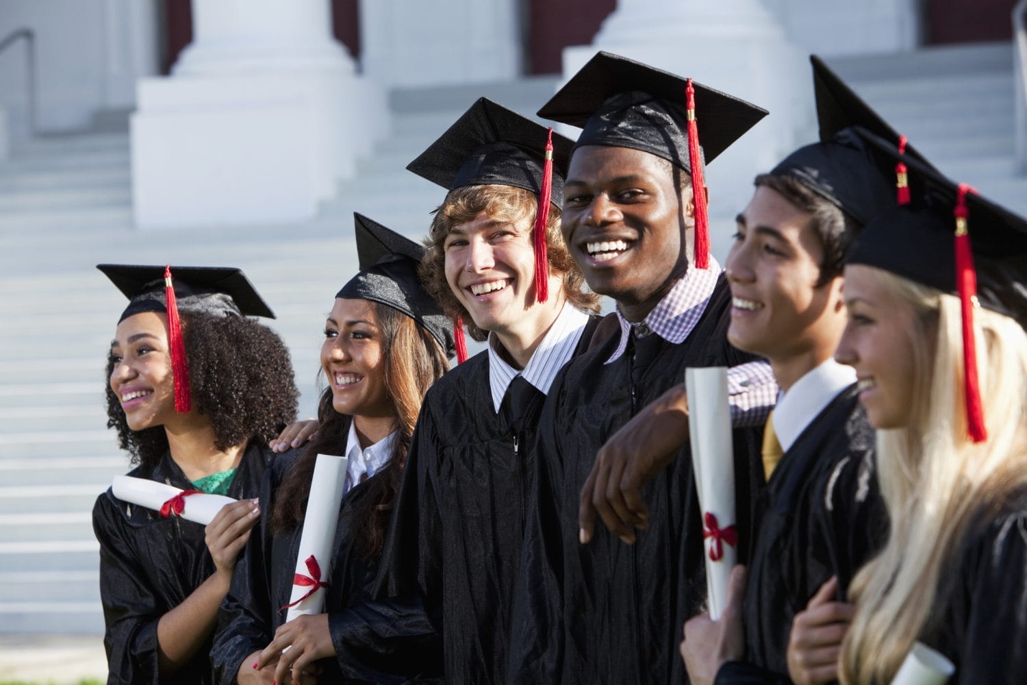 Six diverse students in graduation caps and gowns stand in a row, smiling and holding diplomas, with large white steps and columns in the background, suggesting a graduation ceremony.