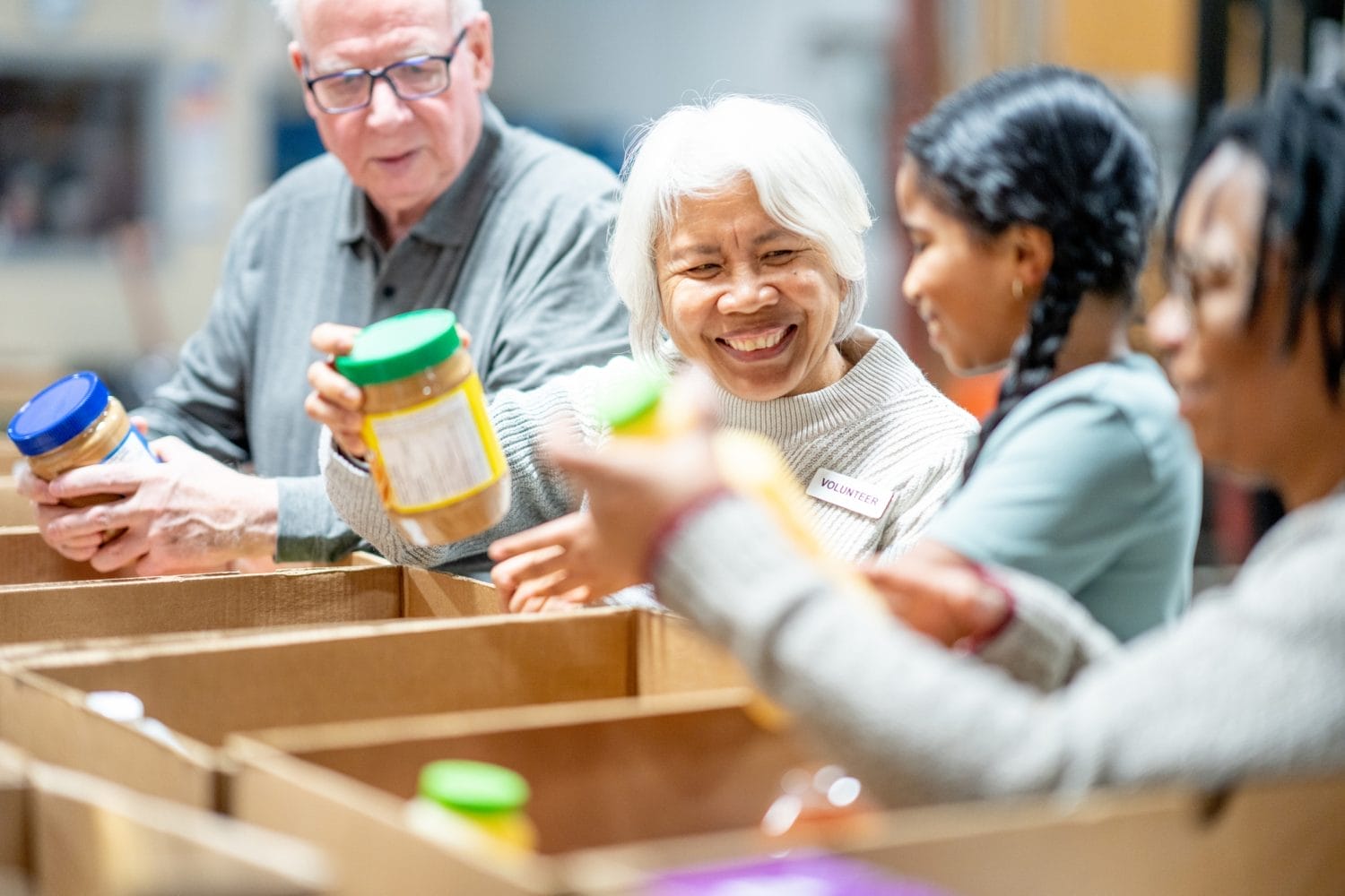 A group of volunteers smiles as they sort jars of food into cardboard boxes at a community food bank.