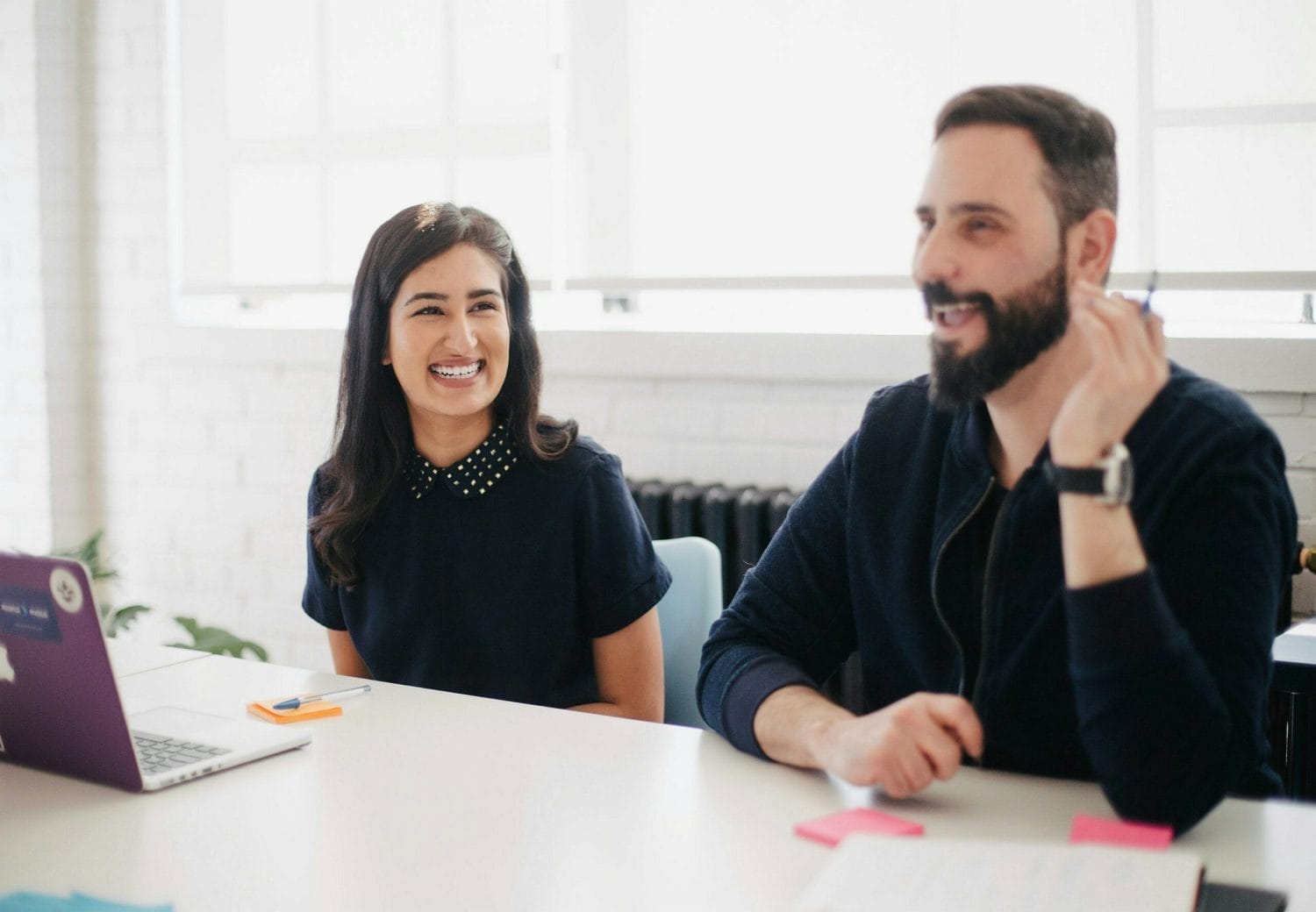 Two people sit at a table in a bright office, smiling and talking. A woman with long dark hair is next to a man with a beard. A laptop, notepad, and sticky notes are on the table in front of them.