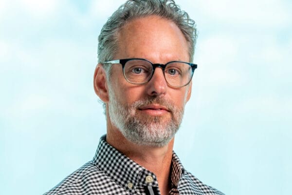 A middle-aged man with gray hair, a short beard, and glasses is wearing a black-and-white checkered shirt, posing against a light blue background.