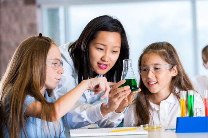 A teacher and two young students wearing safety goggles examine a beaker filled with green liquid in a science classroom. The teacher and students look engaged and curious, with notebooks and colorful test tubes on the table.