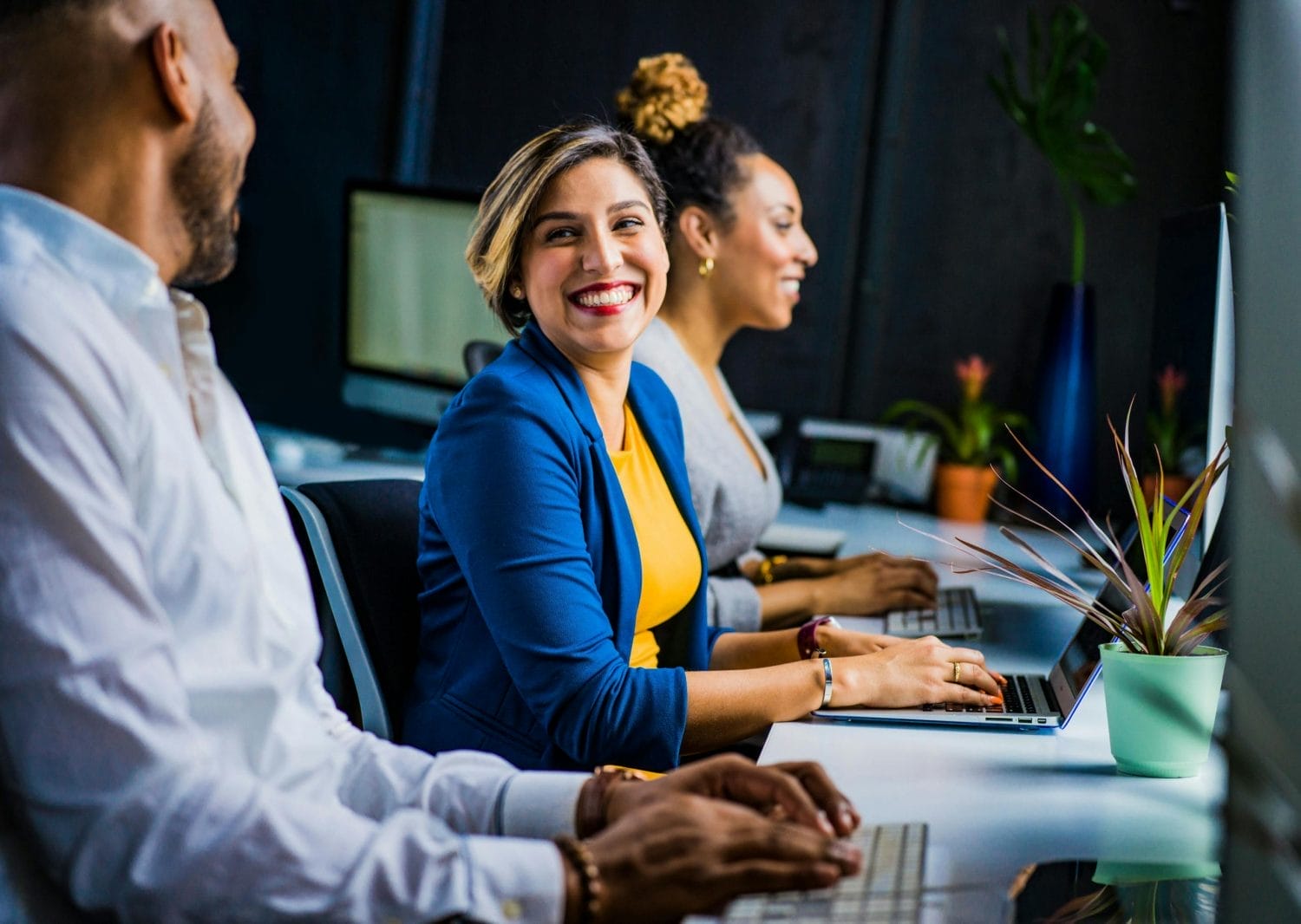 Three people sit at a desk with laptops in an office setting. A woman in a blue blazer and yellow shirt smiles at a colleague, while another woman works in the background. There are plants and computers on the desk.