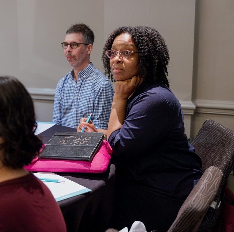 Four adults sit around a conference table with notebooks, papers, and water glasses, attentively listening to a discussion on nonprofit technology strategy. One woman rests her chin on her hand, while another sits in the foreground with eyes closed.