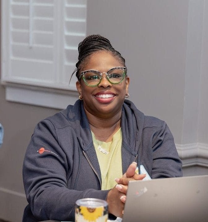 A woman wearing glasses and a dark hoodie is sitting at a table with a laptop and sunflower-patterned tumbler, smiling at the camera during a nonprofit case study meeting or workshop. Other people and papers are visible around her.