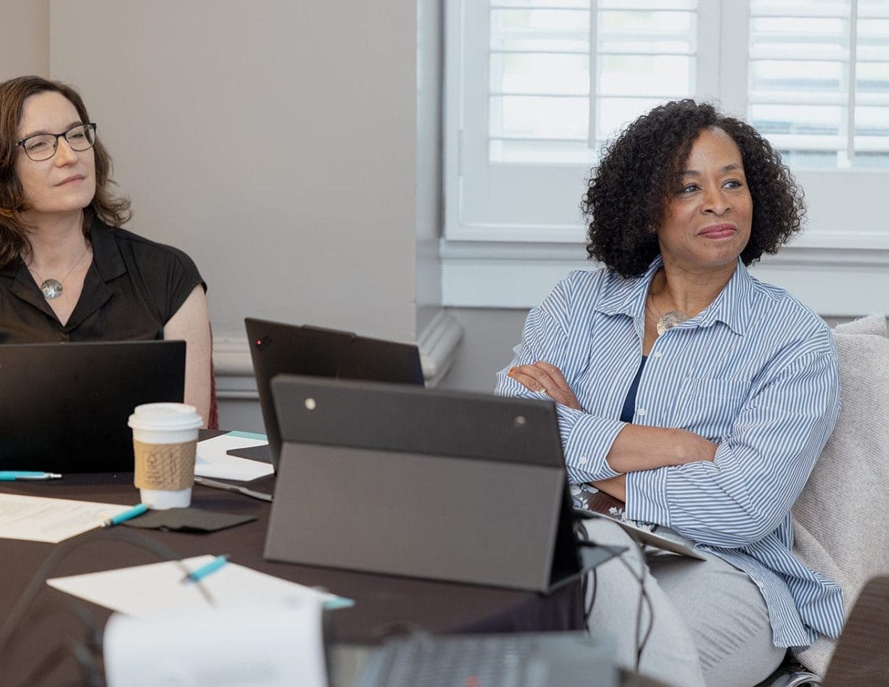 Two women sit at a table with laptops, notebooks, and coffee, attentively listening during a nonprofit or higher ed technology consulting meeting in a bright room with a window and shutters in the background.