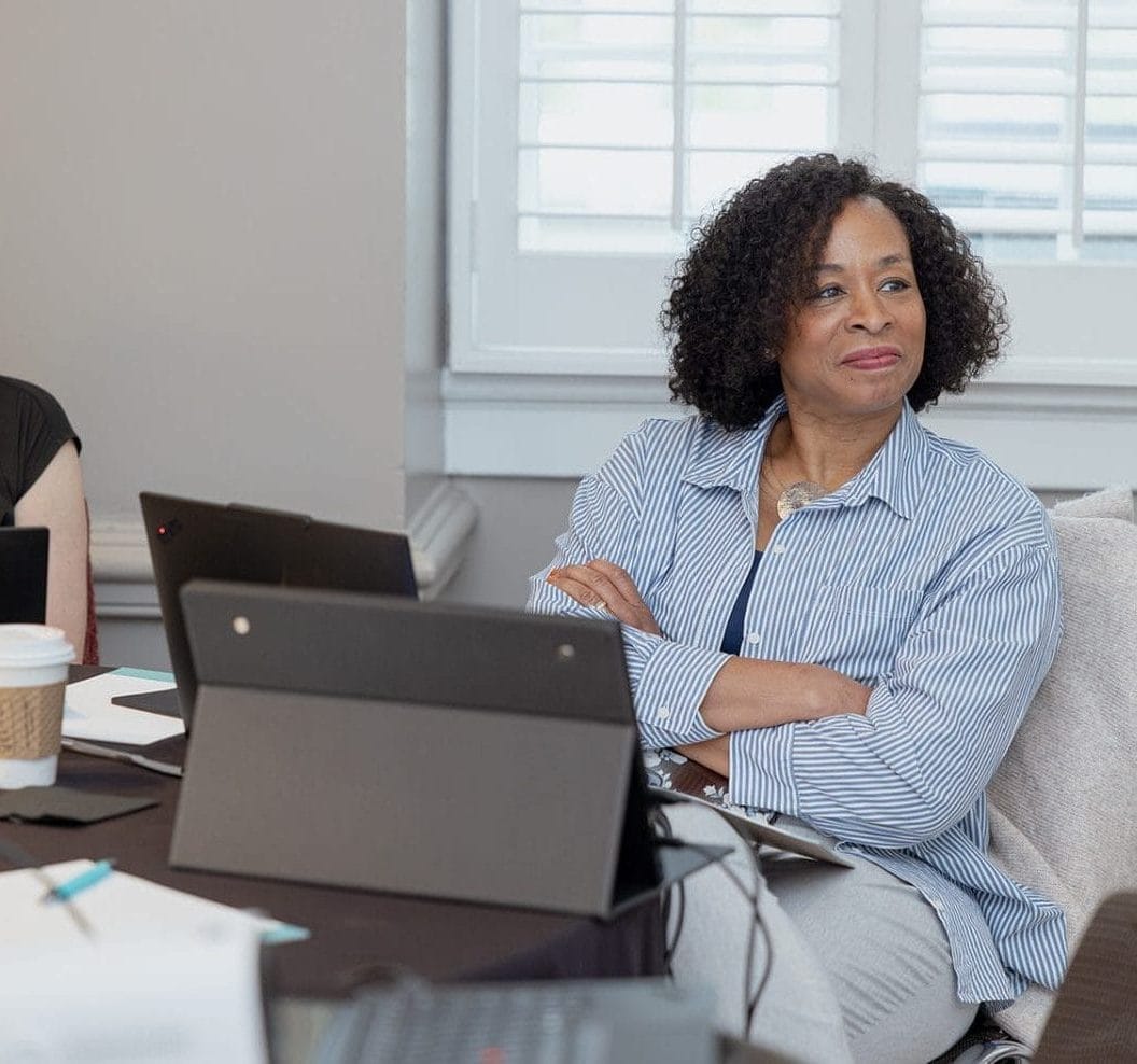 Two women sit at a table with laptops, notebooks, and coffee, attentively listening during a nonprofit or higher ed technology consulting meeting in a bright room with a window and shutters in the background.