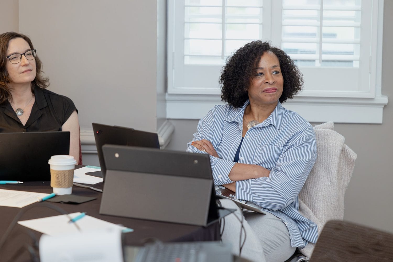 Two women sit at a table with laptops, notebooks, and coffee, attentively listening during a nonprofit or higher ed technology consulting meeting in a bright room with a window and shutters in the background.