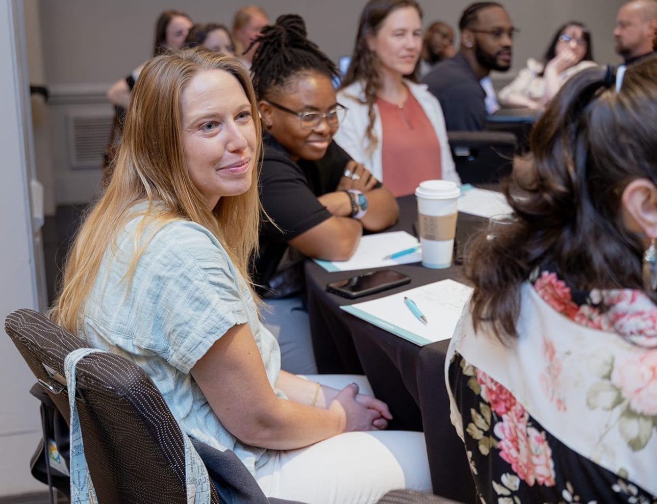 A woman with long blonde hair sits at a table in a conference room, smiling at the camera. Other people, including a Blackbaud consultant, are seated nearby with notebooks and coffee cups, engaged in discussion.