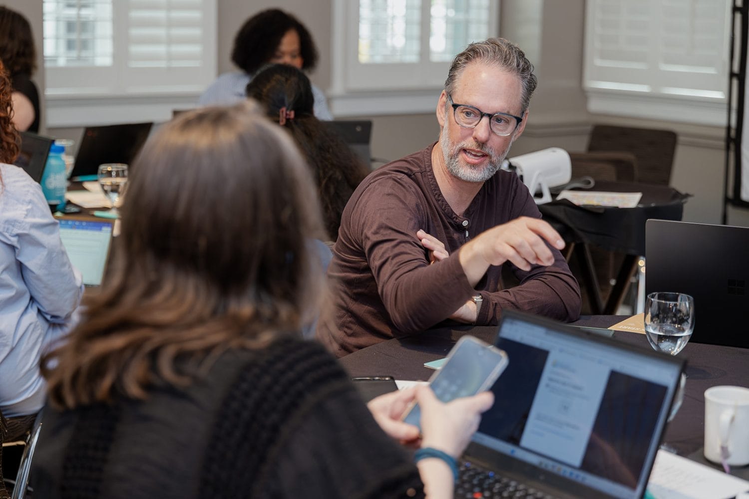 A man with glasses gestures while discussing a nonprofit case study with a woman holding a smartphone at a table filled with laptops and papers in a meeting room. Other people are working in the background.