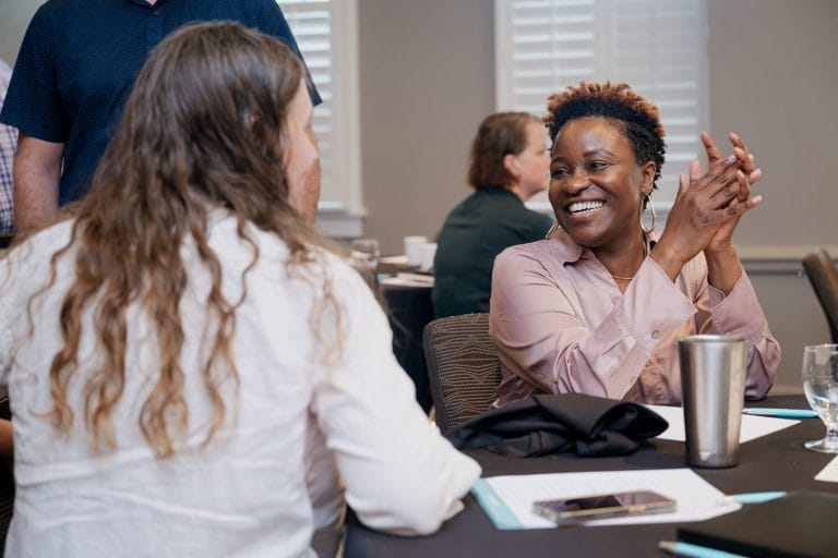 Two people sit at a table talking and smiling during a nonprofit case study workshop. One woman is clapping and laughing while the other listens. Papers, a tumbler, and glasses are on the table, with other participants in the background.