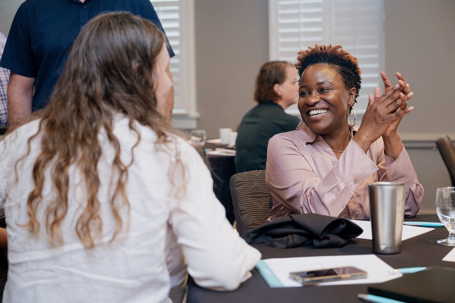 Two people sit at a table talking and smiling during a nonprofit case study workshop. One woman is clapping and laughing while the other listens. Papers, a tumbler, and glasses are on the table, with other participants in the background.