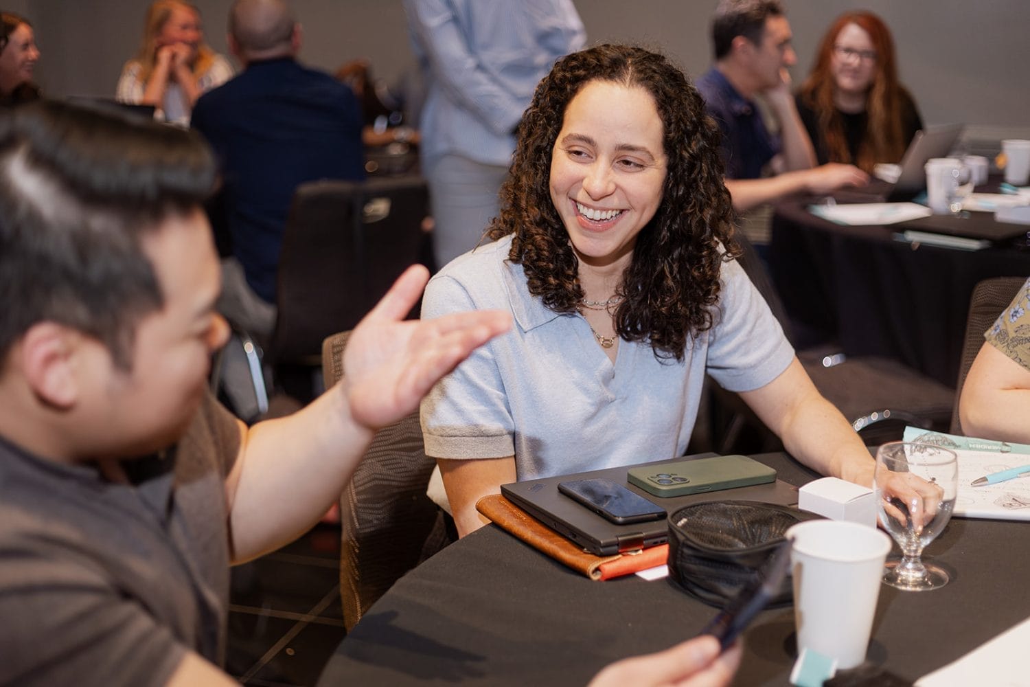 A woman with curly hair smiles while talking to a man at a round table during a nonprofit case study workshop. Laptops, notebooks, and drinks are on the table, with several other people in the background.