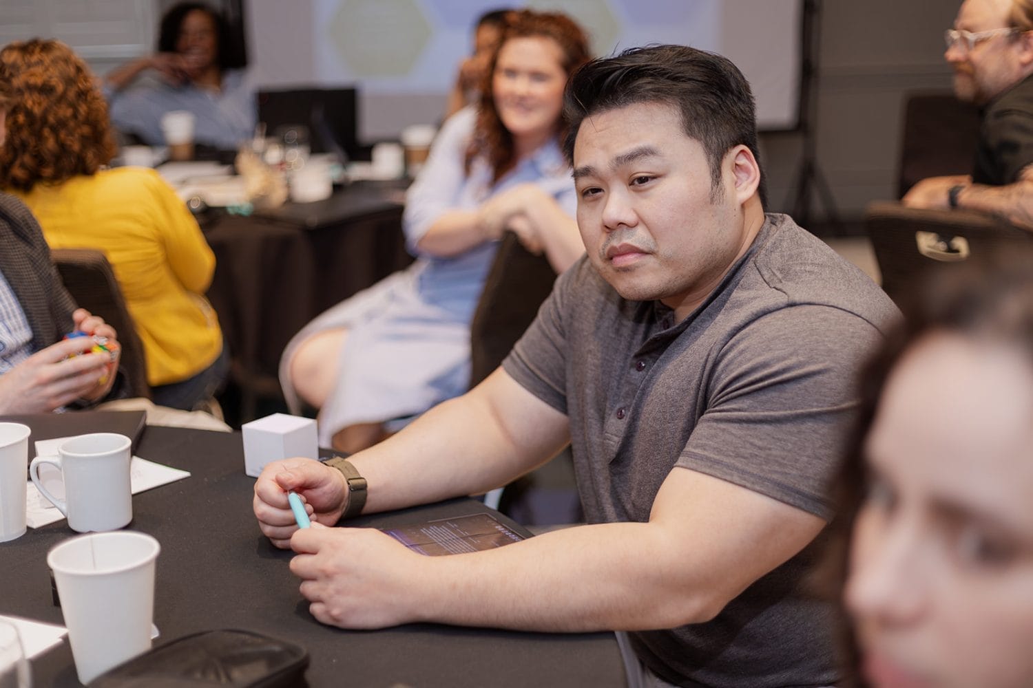 A man sits at a table during a meeting or workshop, holding a pen and looking toward the camera. Several people are seated around tables in the background, engaged in conversation. Cups and papers are on the table.