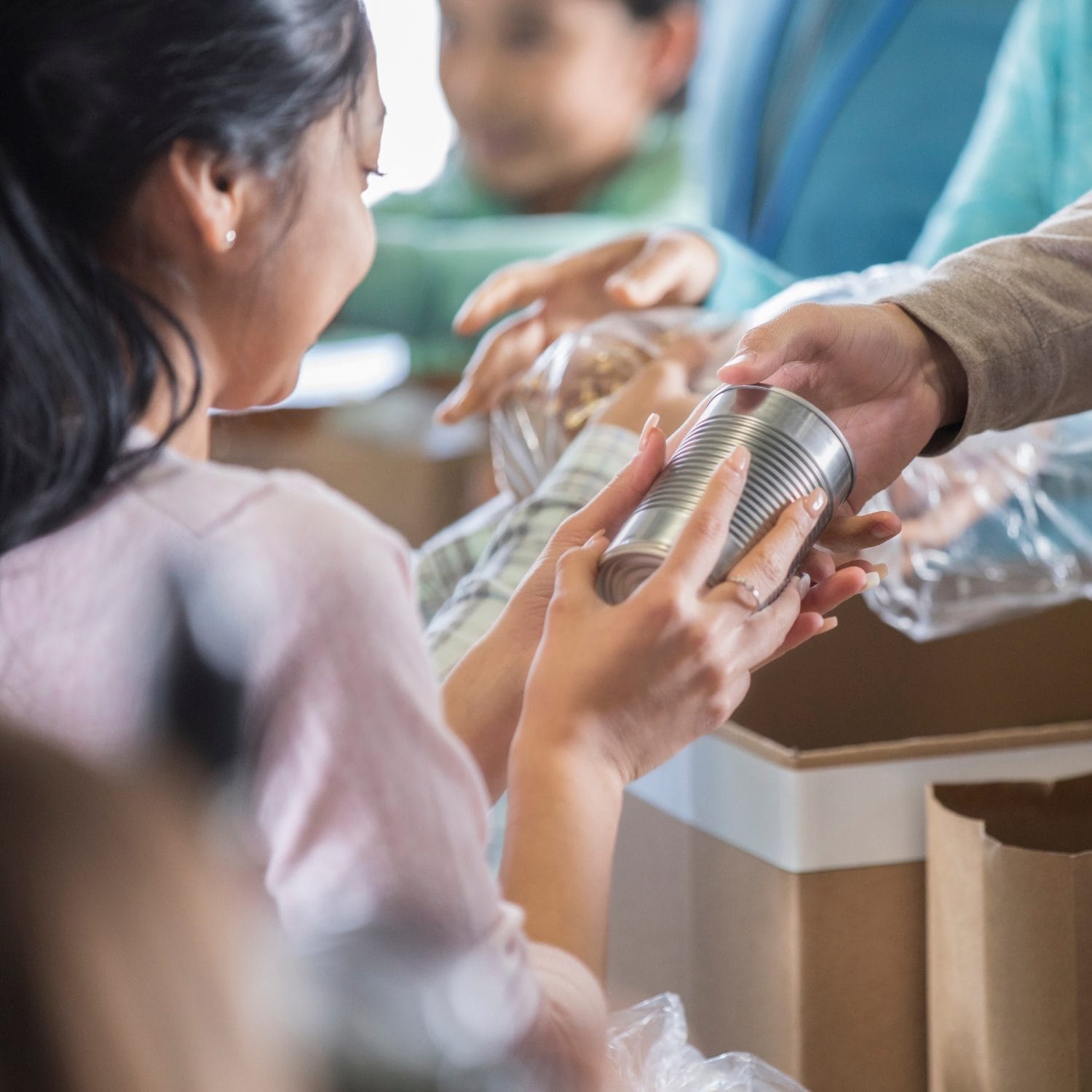 A person hands a tin can to a smiling woman at a food drive or donation center, with boxes and other volunteers visible in the background.