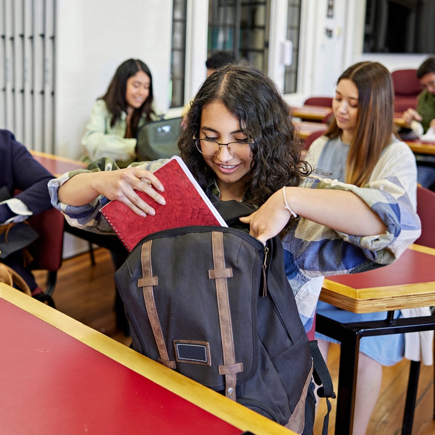 A student with curly hair and glasses puts a red notebook into a black backpack at her desk in a classroom. Other students sit at desks in the background, some smiling and engaged.