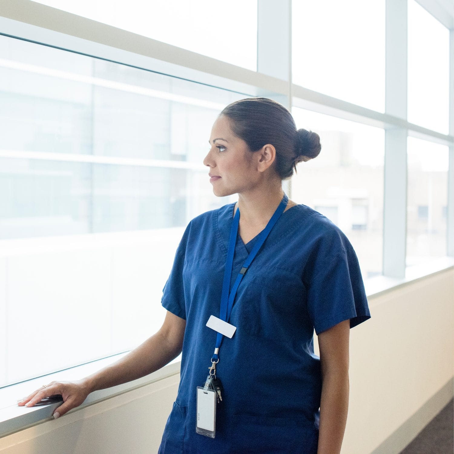 A person in blue medical scrubs and a lanyard stands by a large window, looking outside with a thoughtful expression in a bright, modern building.