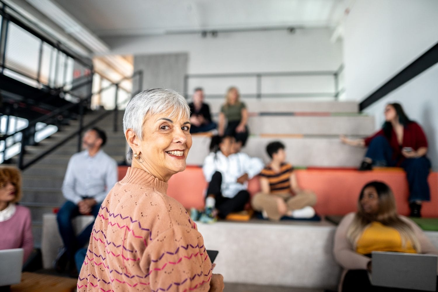 An older woman with short gray hair smiles at the camera in a modern, tiered seating area, while a group of diverse people in casual clothes sit and talk in the background.