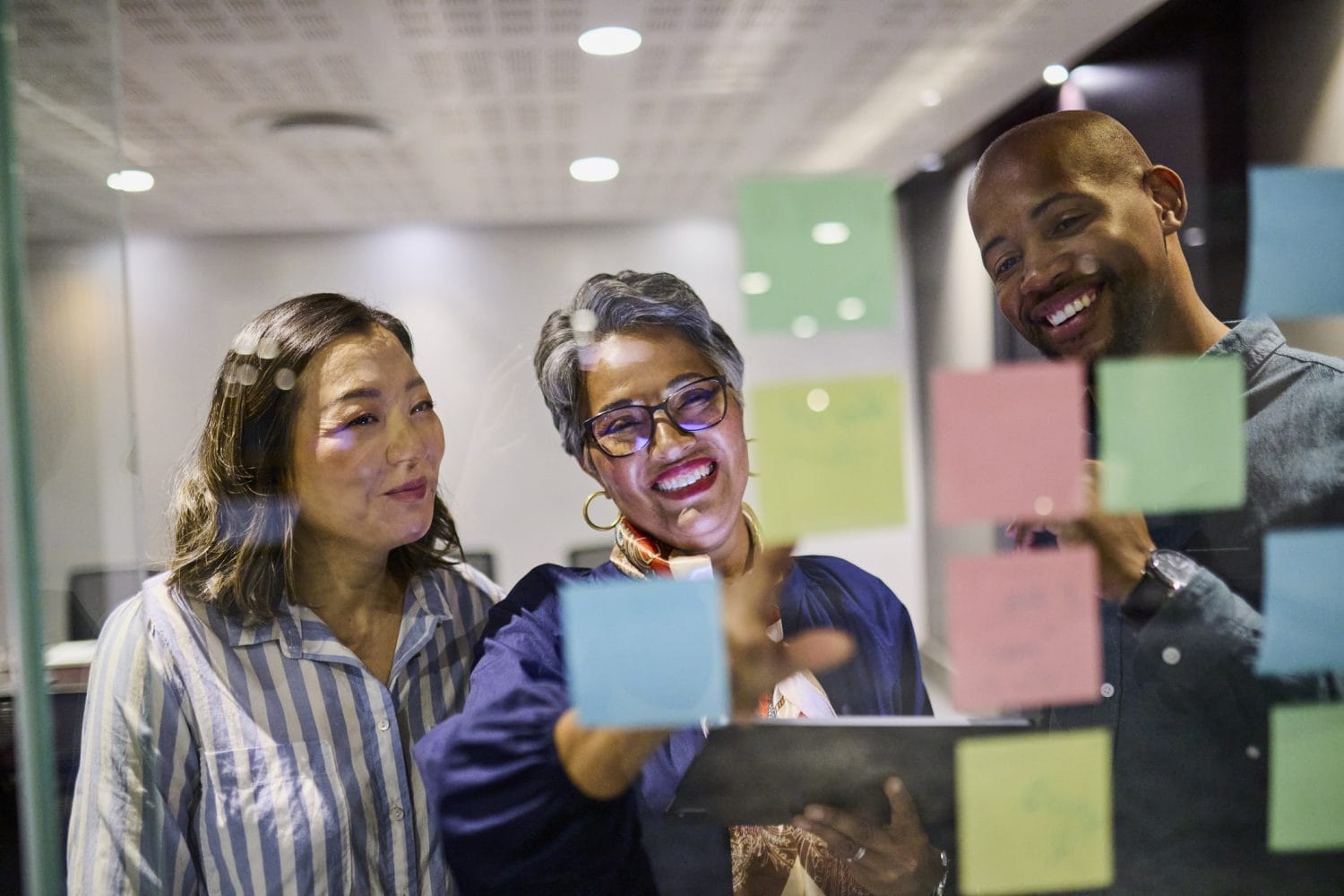 Three people stand in front of a glass wall covered with colorful sticky notes, smiling and discussing ideas. One person points at a note while holding a tablet, reflecting a collaborative nonprofit and higher ed technology consulting environment.