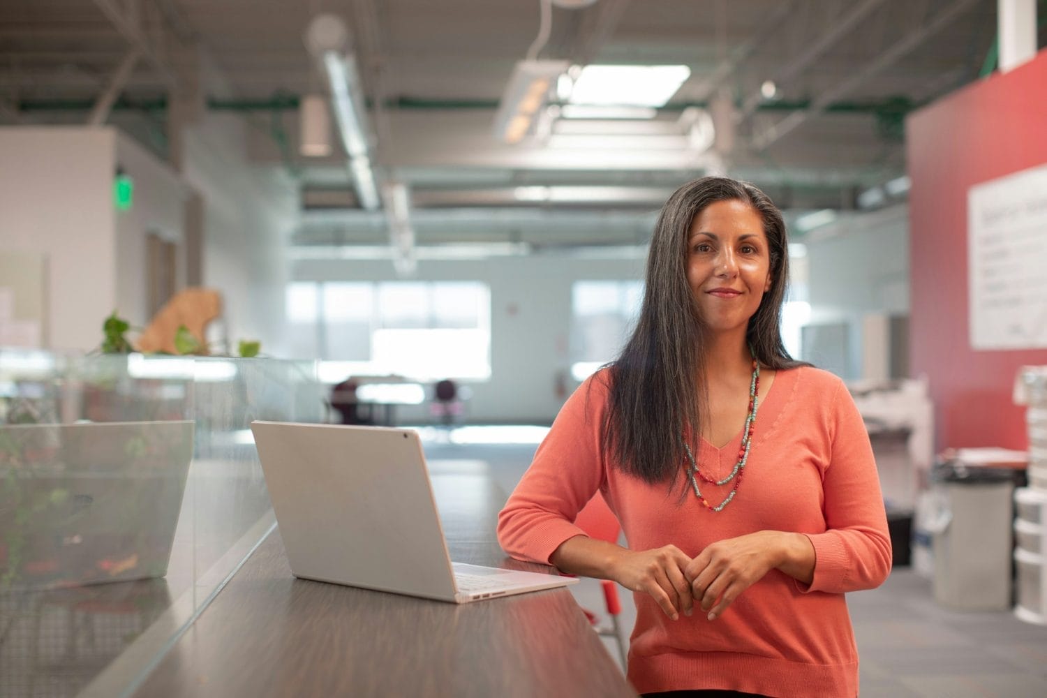 A woman with long dark hair, wearing a peach sweater and colorful necklace, stands smiling beside an open laptop in a modern, brightly lit office space—ready to discuss opportunities in nonprofit and higher ed careers.