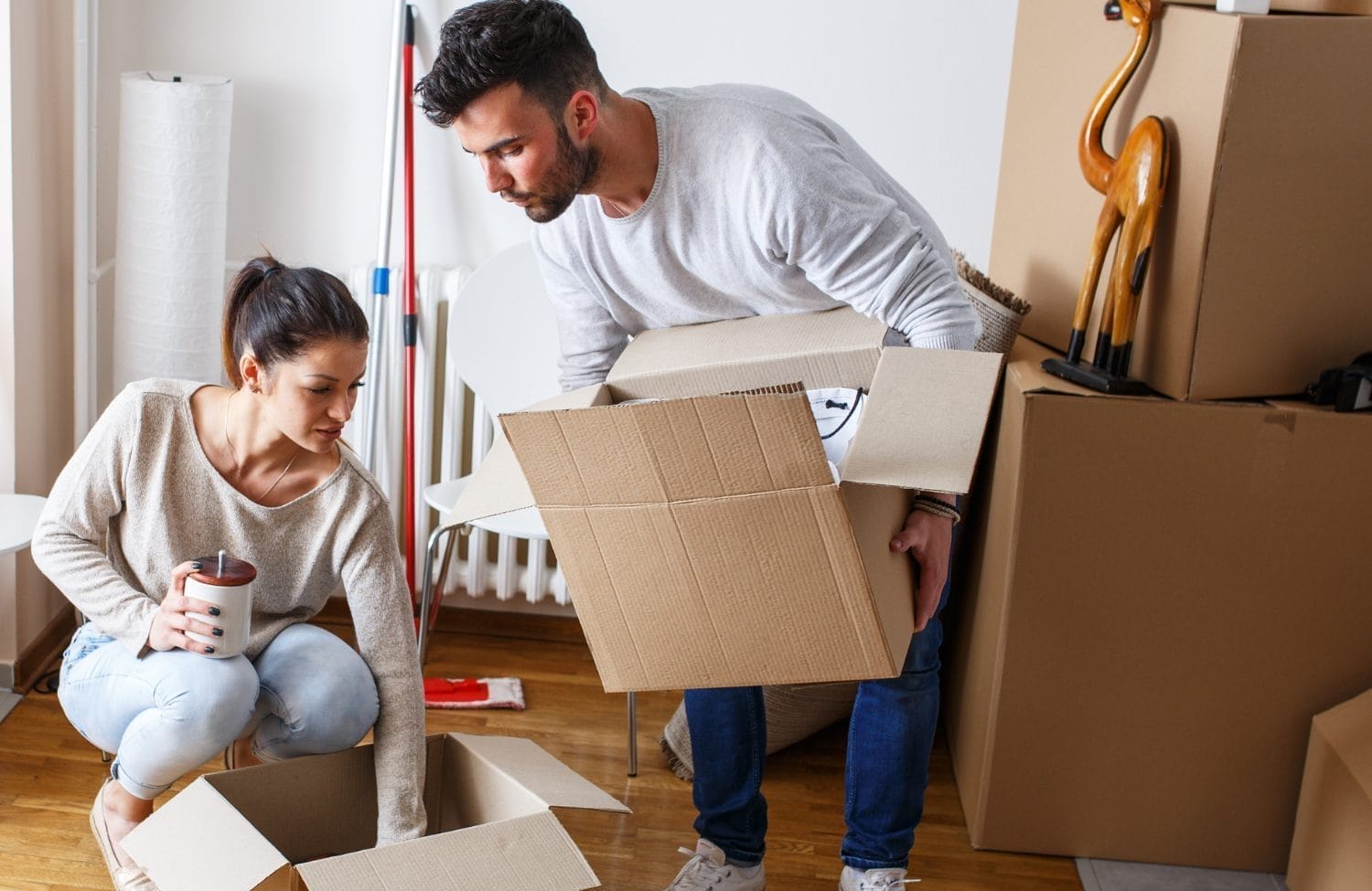 A man and woman packing cardboard boxes in a room with wooden floors; there are various moving boxes, a mop, and a decorative giraffe in the background. The woman holds a mug while the man lifts a box.