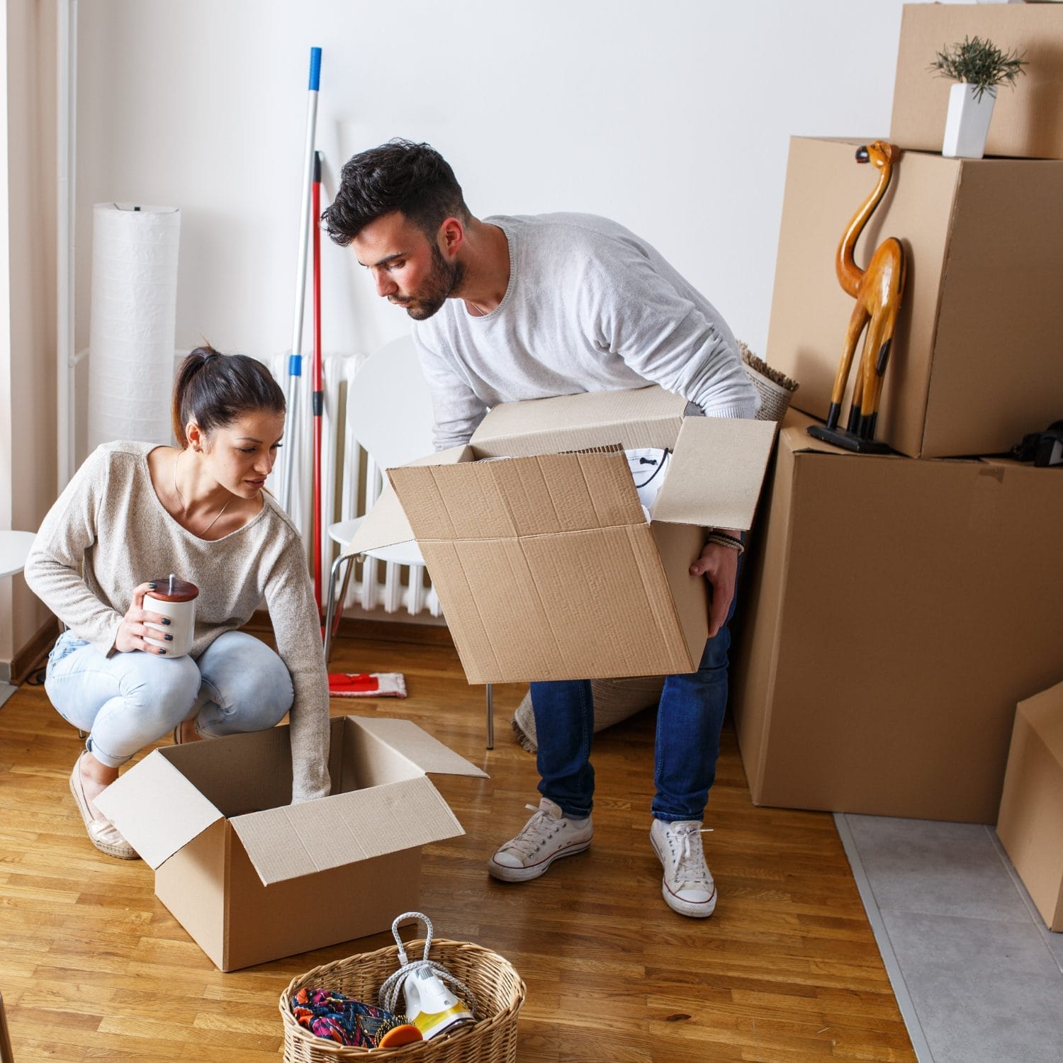 A man and woman packing cardboard boxes in a room with wooden floors; there are various moving boxes, a mop, and a decorative giraffe in the background. The woman holds a mug while the man lifts a box.