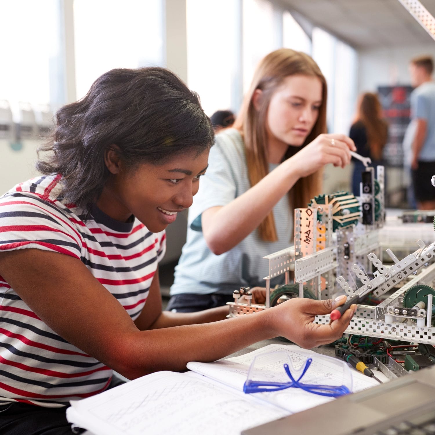 Two young women work together on building a metal robotics project, surrounded by tools and notebooks, in a bright classroom or lab setting. One smiles while adjusting a component, the other focuses on assembling parts.
