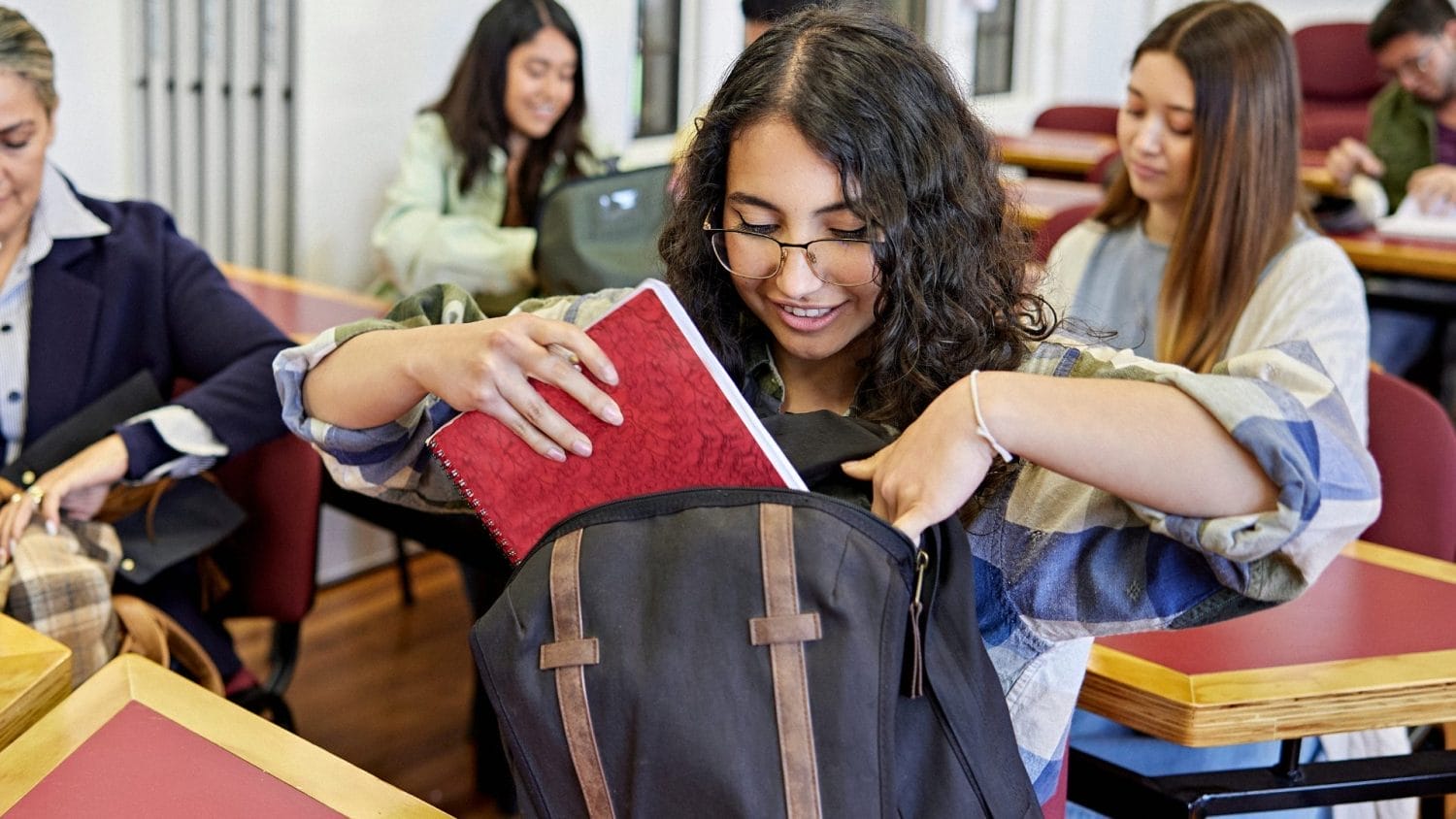 A student with curly hair and glasses smiles while placing a red notebook into her backpack in a classroom, with other students sitting at desks in the background.