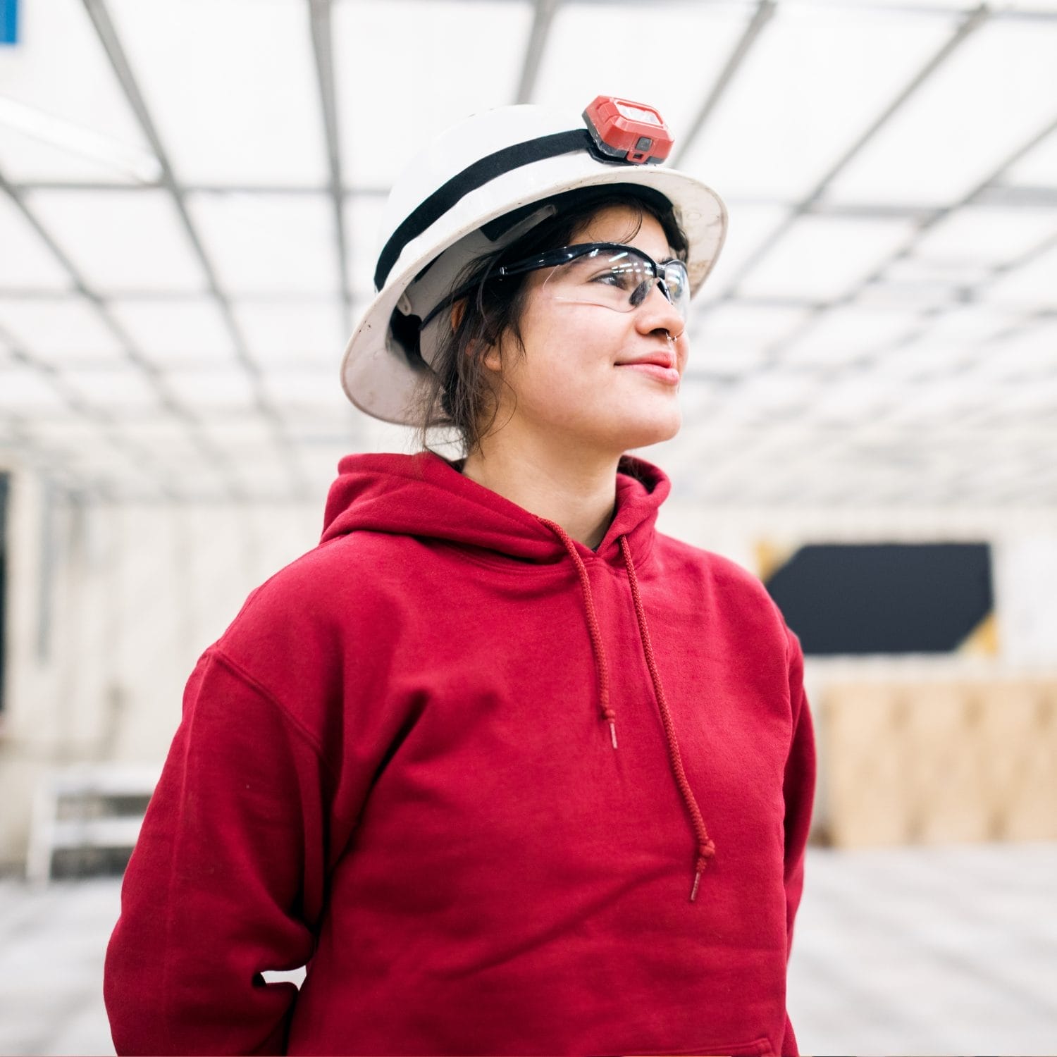 A young person wearing a white hard hat, safety glasses, and a red hoodie stands indoors, looking up and smiling confidently in a bright, industrial space.