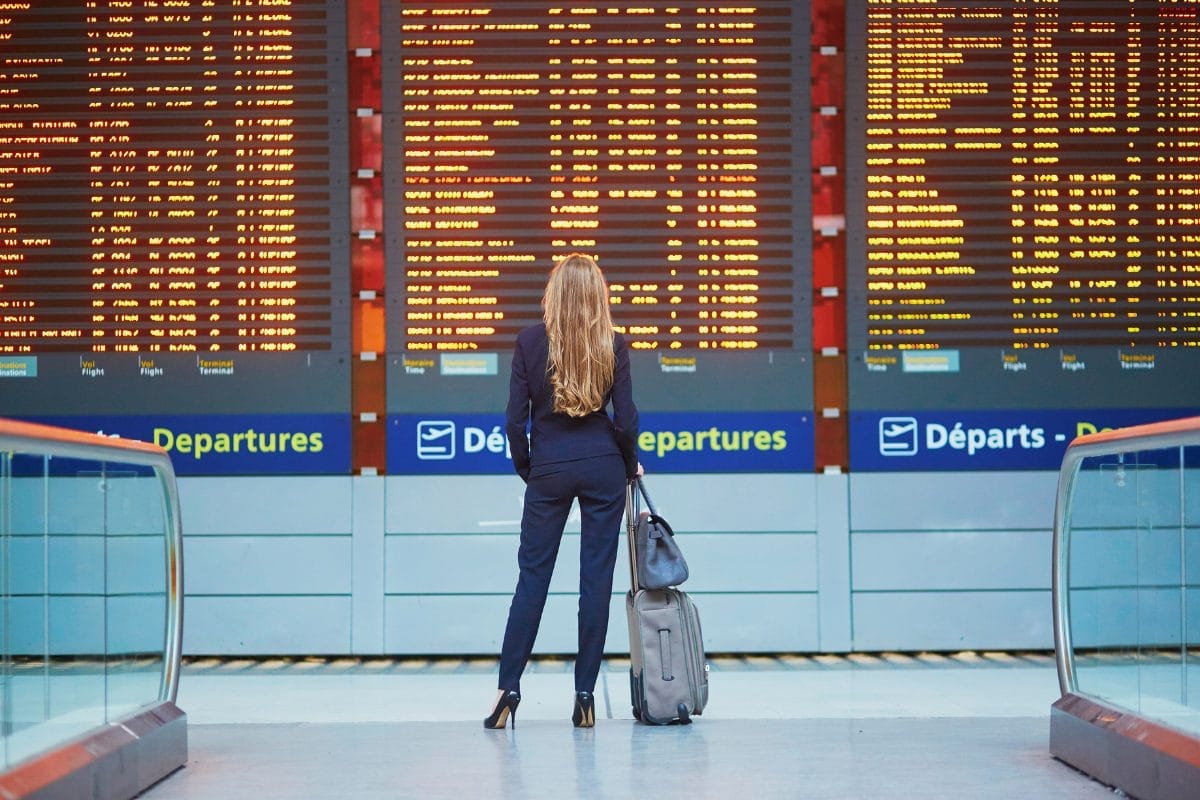 A woman with long hair stands in front of a large departures board at an airport, holding the handle of her rolling suitcase and looking up at the listed flight information.