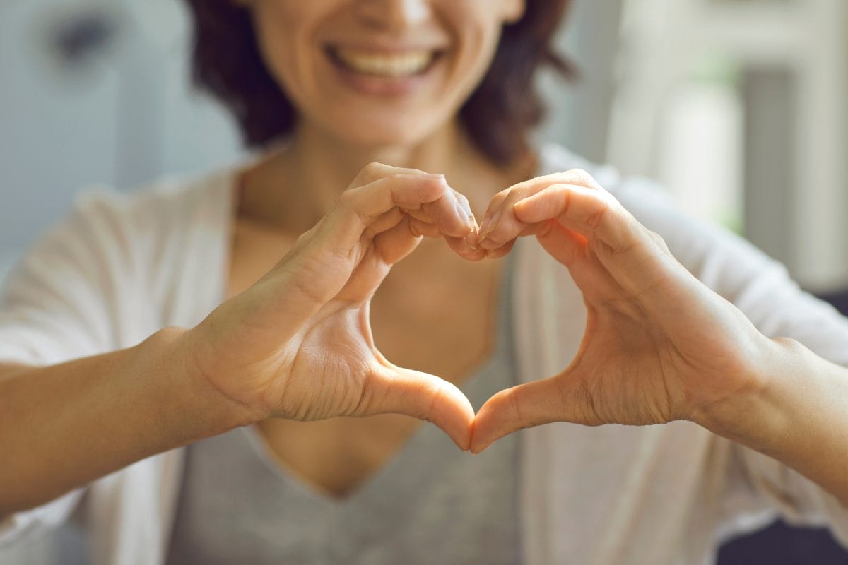 A woman smiles while forming a heart shape with her hands in front of her chest. The focus is on her hands and the heart gesture, with her face partially visible and out of focus in the background.
