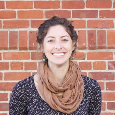 A woman with short curly brown hair, wearing turquoise earrings, a brown scarf, and a black top with white dots, smiles in front of a red brick wall.