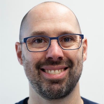 A smiling man with a bald head, a short beard, and mustache, wearing blue rectangular glasses, in front of a plain light background.