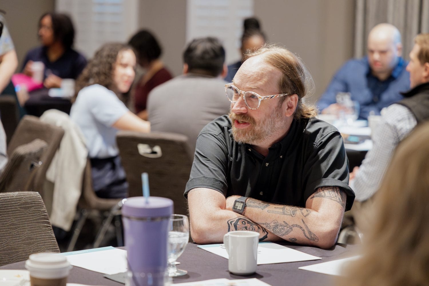 A man with glasses and visible arm tattoos sits at a table with a mug and water glass, engaged in conversation at a busy meeting or workshop with other people in the background.