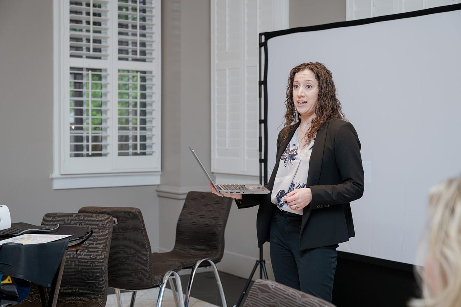 A woman with curly hair in a blazer holds a laptop, presenting on digital tools in a modern conference room with empty chairs and a projector screen behind her.