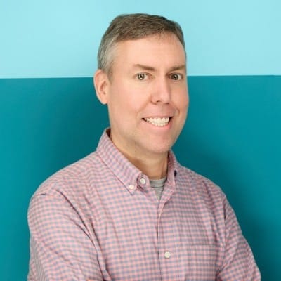 A man with short brown hair, wearing a pink and blue checkered shirt, stands smiling with his arms crossed in front of a two-tone blue background.