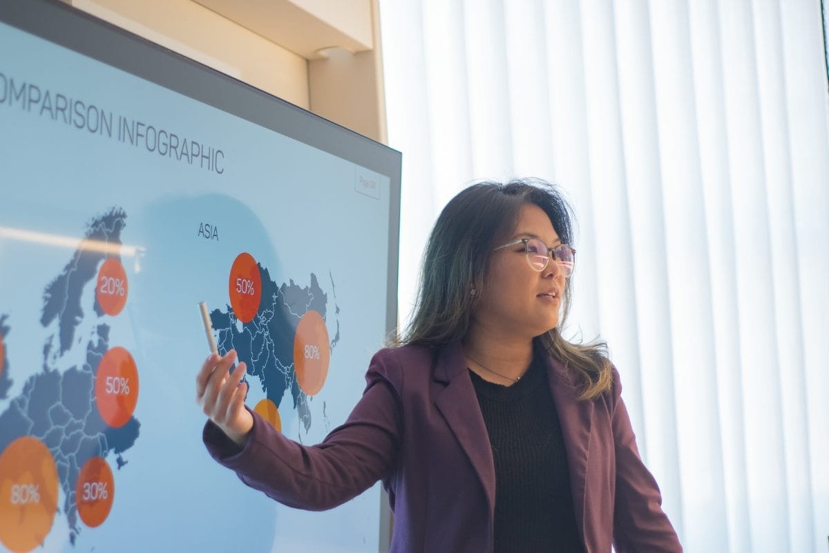 A woman wearing glasses and a purple blazer gives a presentation, pointing at a world map infographic on a screen that displays percentage data for different regions. Light streams in through sheer curtains in the background.