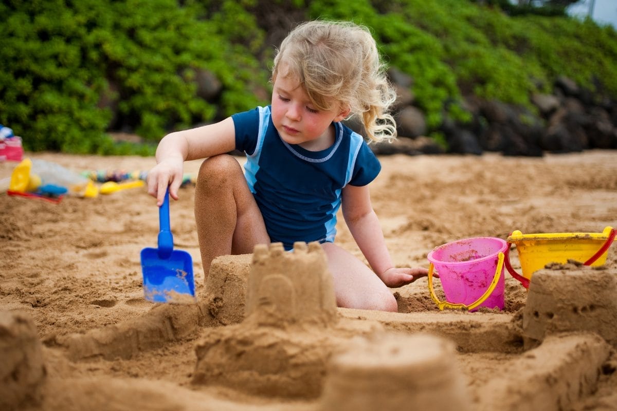 A young child with blonde hair builds a sandcastle on a beach using a blue shovel, with colorful buckets nearby and greenery in the background.