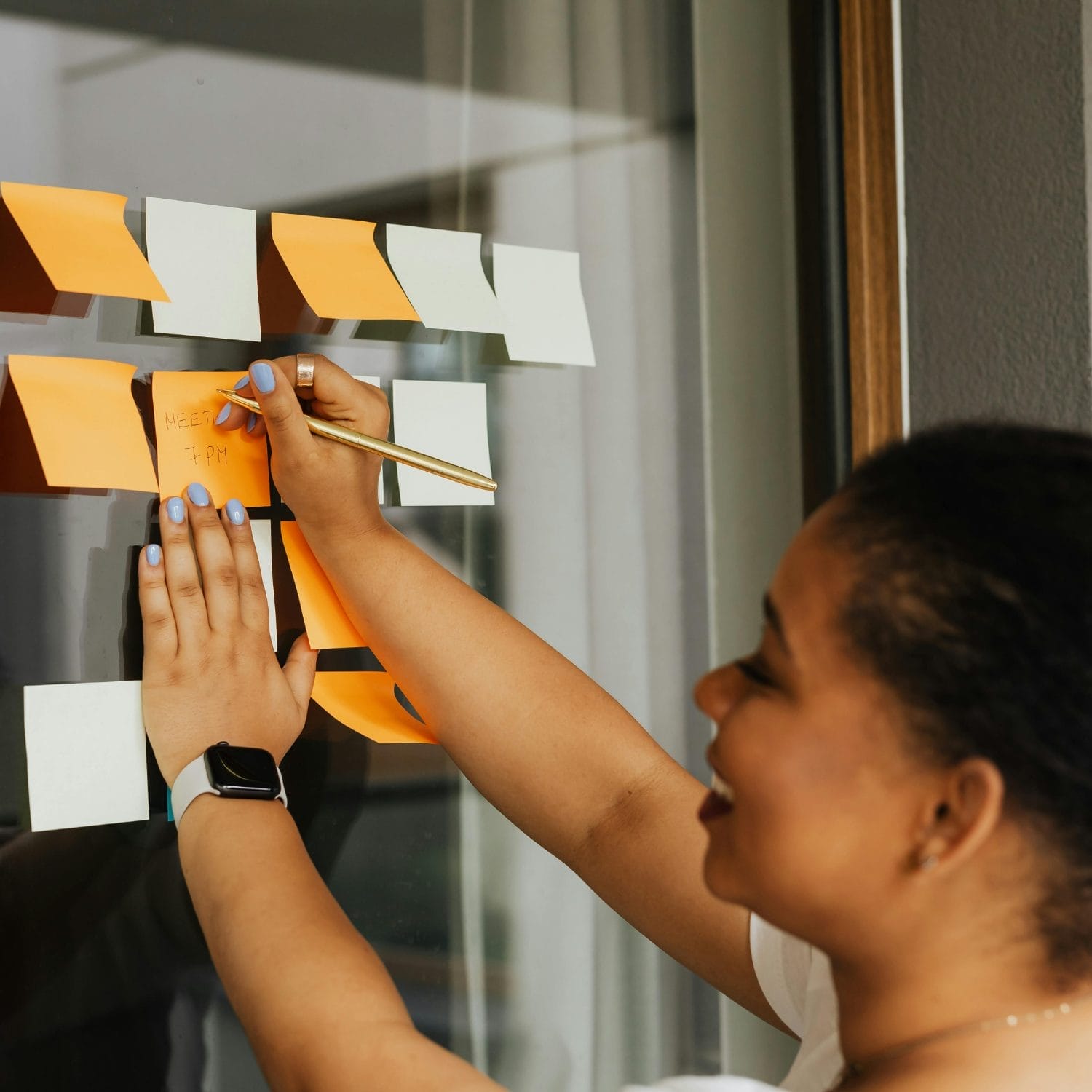 A person smiling while writing on an orange sticky note attached to a glass wall, surrounded by other orange and white sticky notes, with a smartwatch on their wrist.