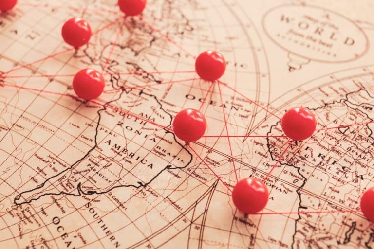 Close-up of a vintage world map with red push pins connected by red string, marking various locations across North America, Europe, and surrounding regions.